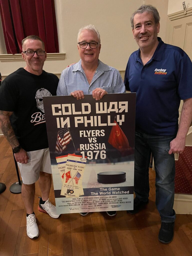 Three men indoors posing with a large vintage poster about Flyers vs. Russia (1976) featuring American and Soviet flags and text from the event promo.